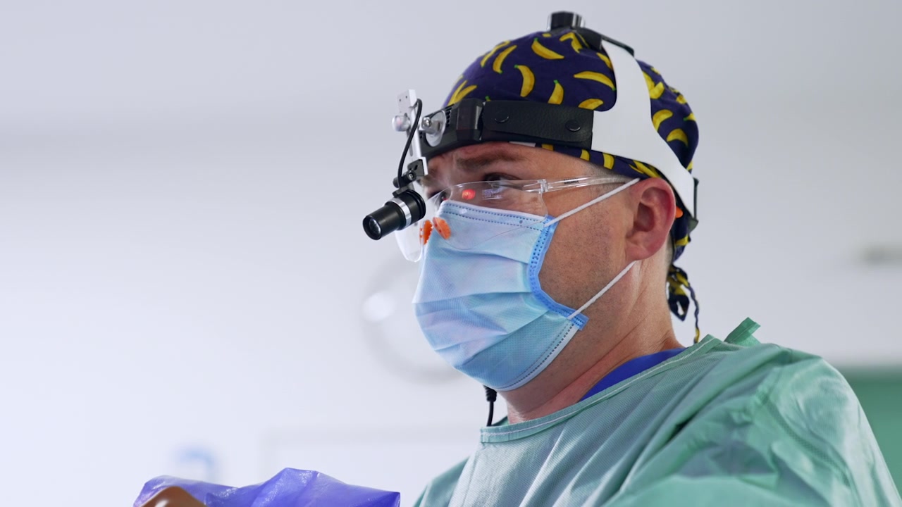 Médico caucasiano usando touca, máscara, óculos de proteção e luz frontal olha para frente. Retrato em close-up de perfil de um cirurgião operando e olhando para a tela