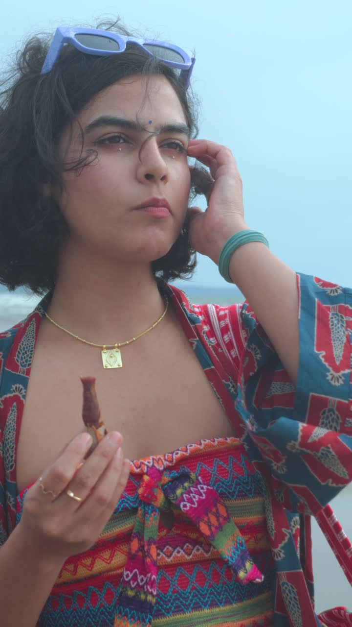 Vertical medium close-up of an Indian woman chewing food at an overcast beach, dressed in a very colourful vibrant modern summer outfit