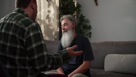 A-happy-man-with-gray-hair-and-a-lush-gray-beard-has-fun-and-rejoices-during-his-communication-with-his-boyfriend-a-brunette-with-stubble-in-a-plaid-shirt-sitting-on-a-gray-sofa-in-a-modern-apartment