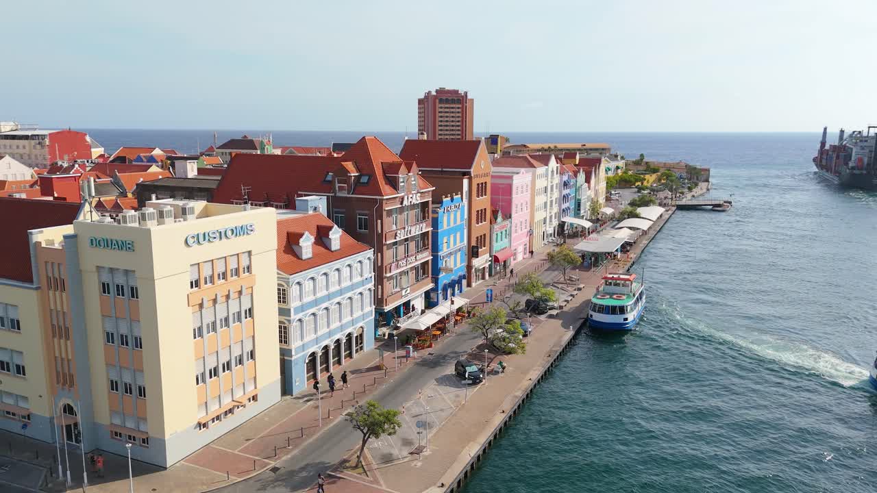 Aerial drone rises above Punda district’s vibrant waterfront buildings along the coast of Curacao, calm sea visible ferry leaves dock and cargo ship heads out to sea