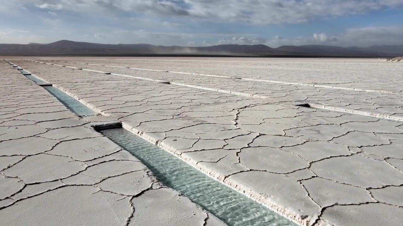 Narrow turquoise brine canals cut through cracked white crust at Salinas Grandes salt flat in Jujuy Province, Argentina, showing lithium-rich extraction patterns under a wide sky.
