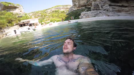 joven nadando de espaldas en una cala oceánica mirando hacia el cielo y los acantilados que rodean el agua clara