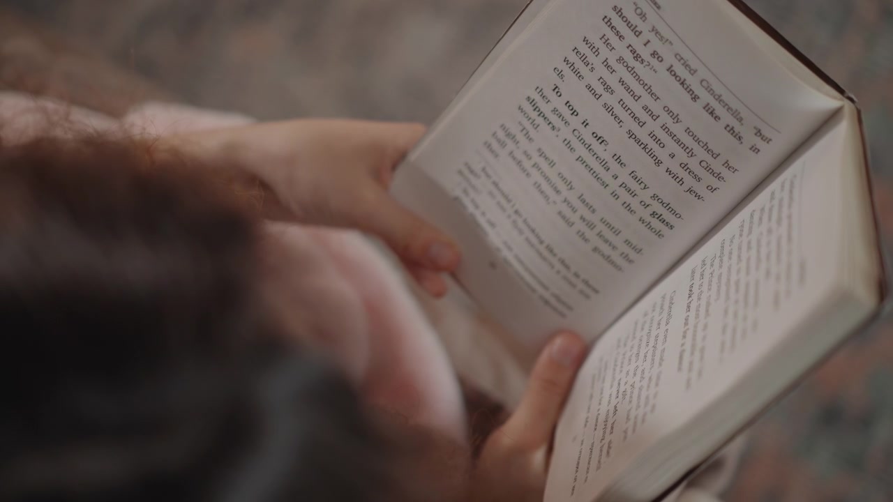 Overhead intimate view of adult hands thumb-through and turn pages of small paperback book, visible page curl and finger motion indicating active reading and page flipping; soft warm indoor lighting