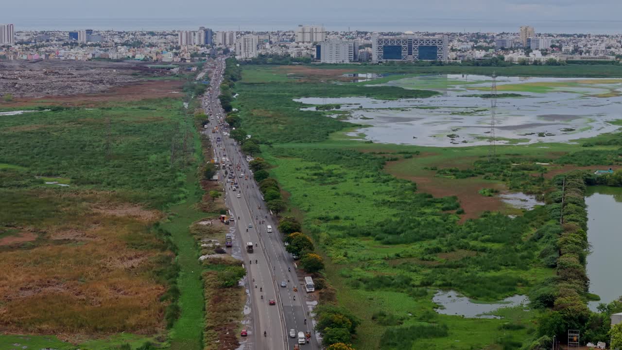 Pan view of straight busy road joining cities with green, flooded field on either side and cityscape at background. Drone view.