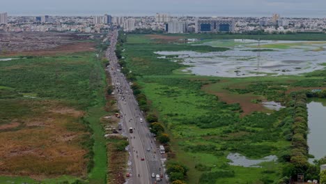 pan view of straight busy road joining cities with green, flooded field on either side and cityscape at background. drone view.