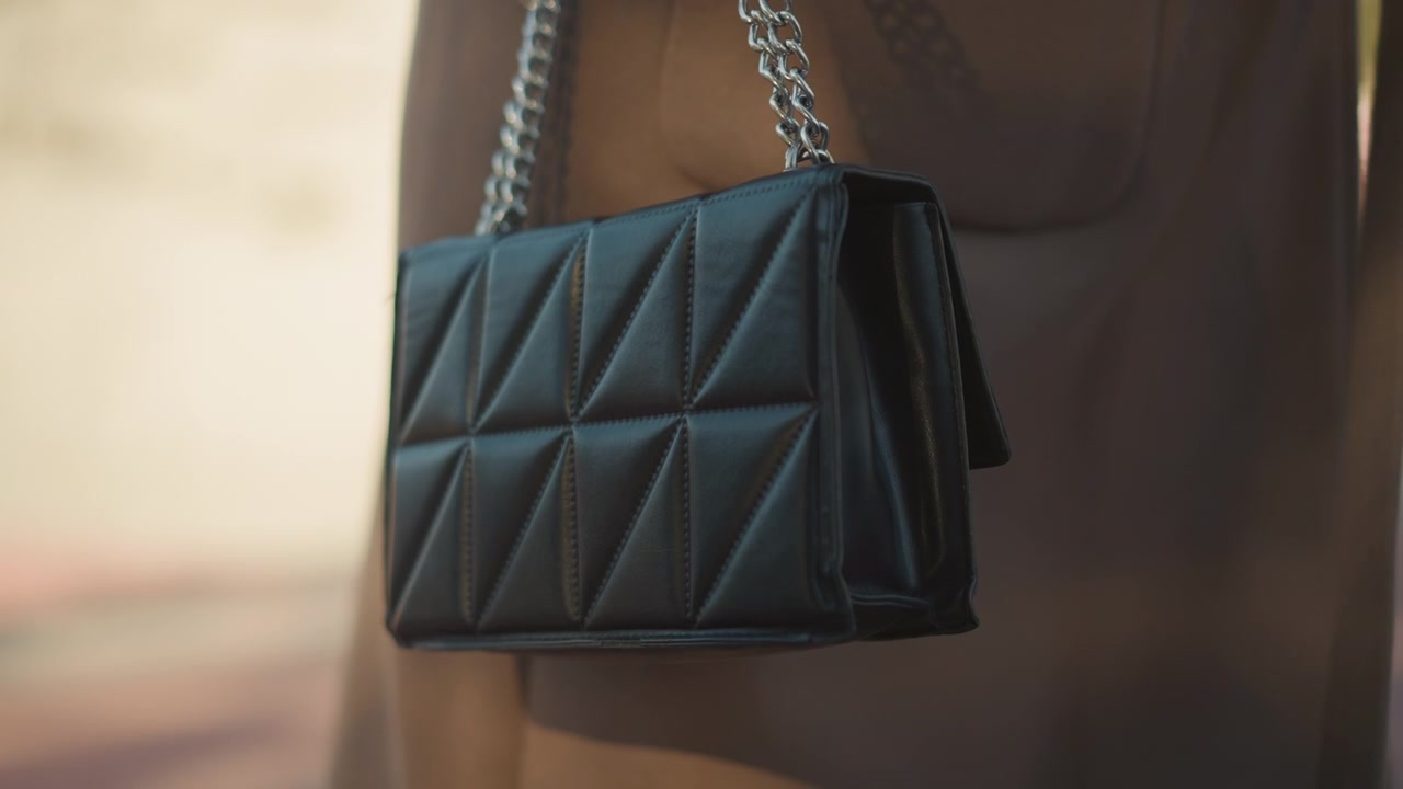 Primo piano di una borsa in pelle trapuntata nera con tracolla a catena argentata che pende dal braccio. La borsa presenta dettagli di cuciture geometriche e riflessi di luce soffusa su uno sfondo sfocato.