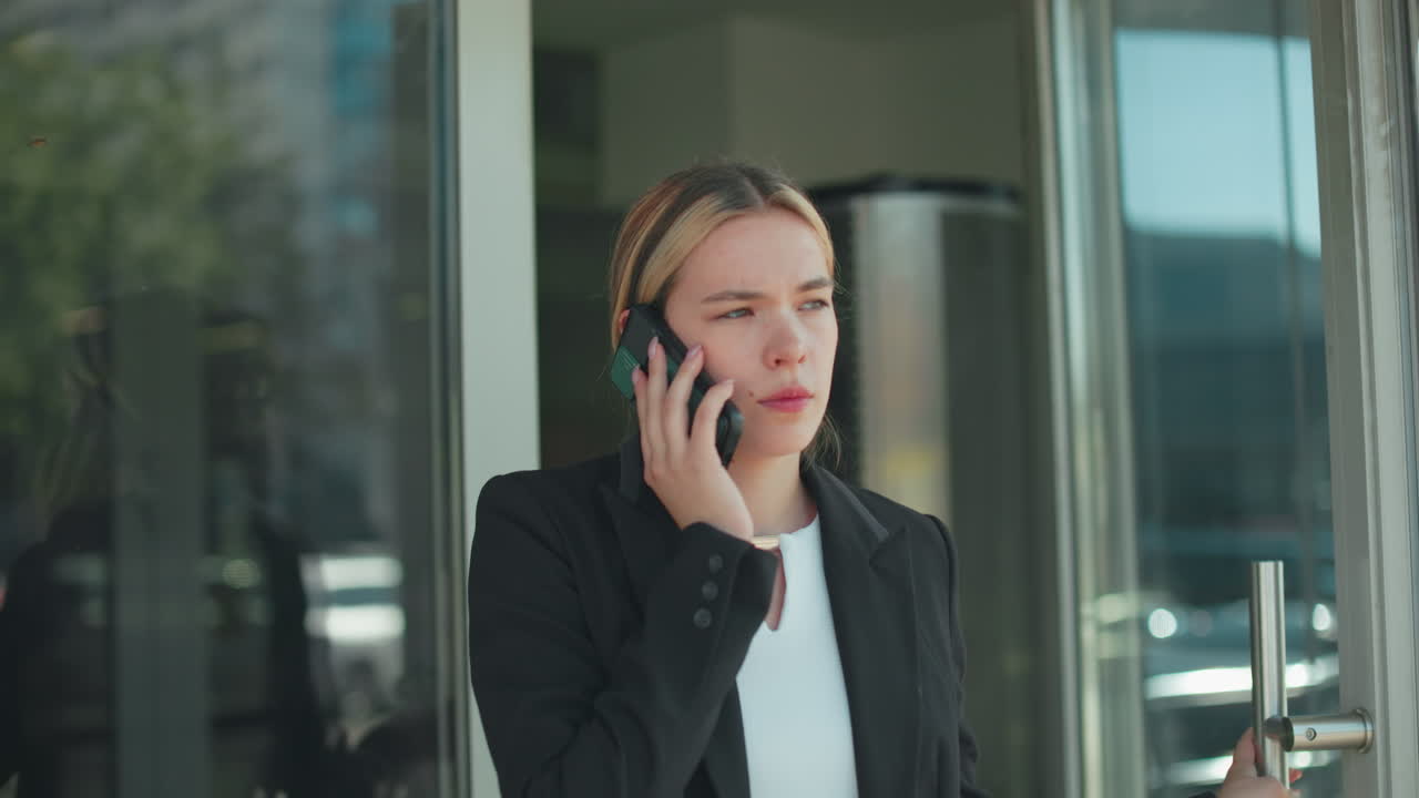 Young girl exits conference hall while talking on phone with client, thoughtful expression on face, walking with sunlight reflecting on her face, urban cityscape visible through glass