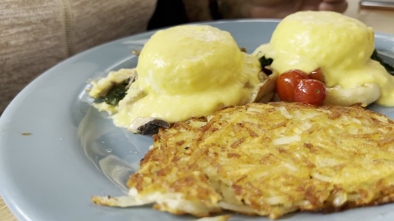 Two poached eggs with hollandaise sauce are served on toasted english muffins with sauteed spinach, roasted cherry tomatoes, and crispy hash browns for a complete satisfying brunch, slow motion reveal