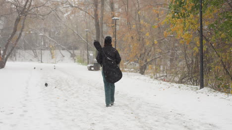 back view of individual in black beanie and hoodie with hands in pocket carrying guitar in pack walking in snowfall along snowy path with bench, streetlights, trees, and distant pigeons in background