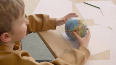 vue de dessus d'un enfant blond jouant avec un globe assis à une table où sont des dessins de la terre