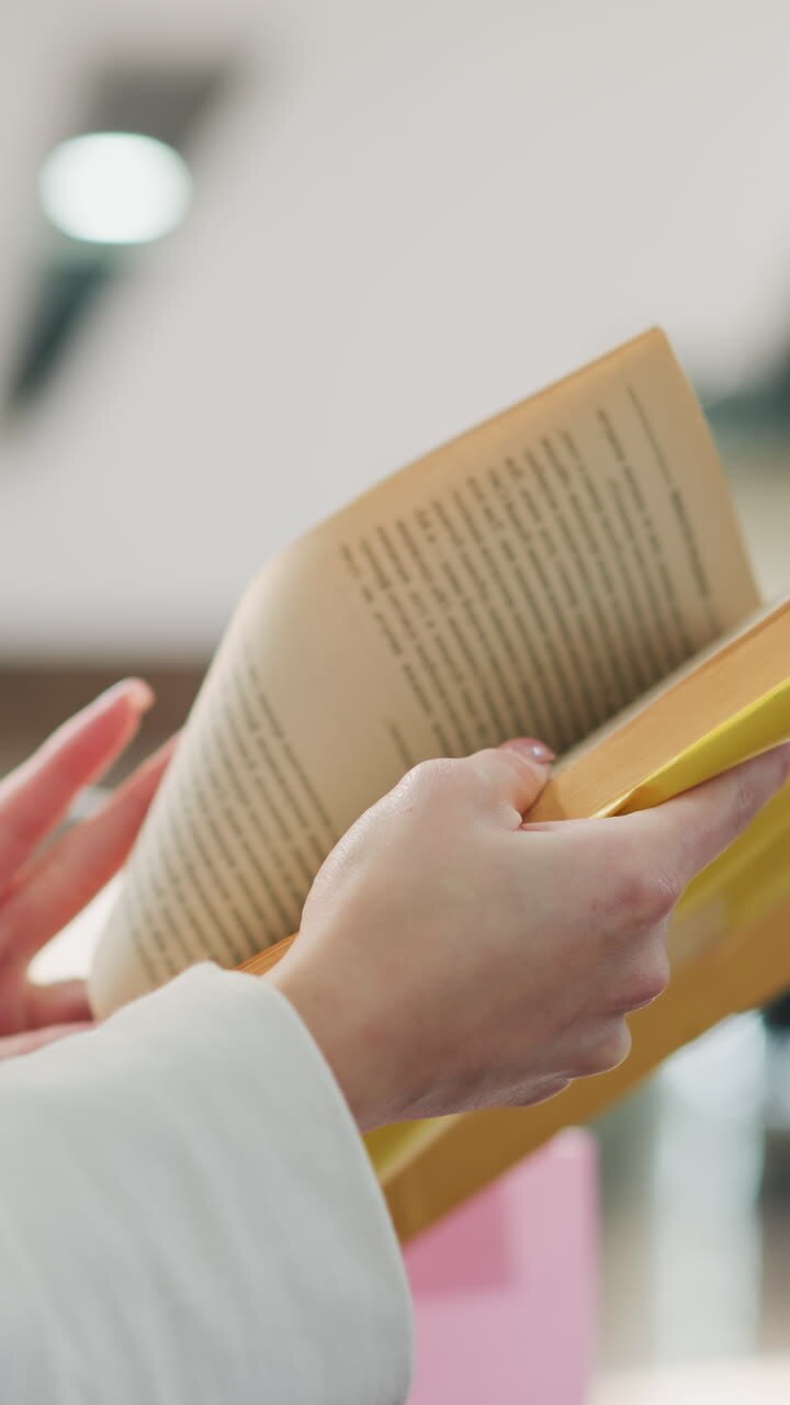Close-up of hands flipping through a yellow-covered book, showcasing elegant gestures and delicate details, set in a softly lit indoor environment with a blurred modern background