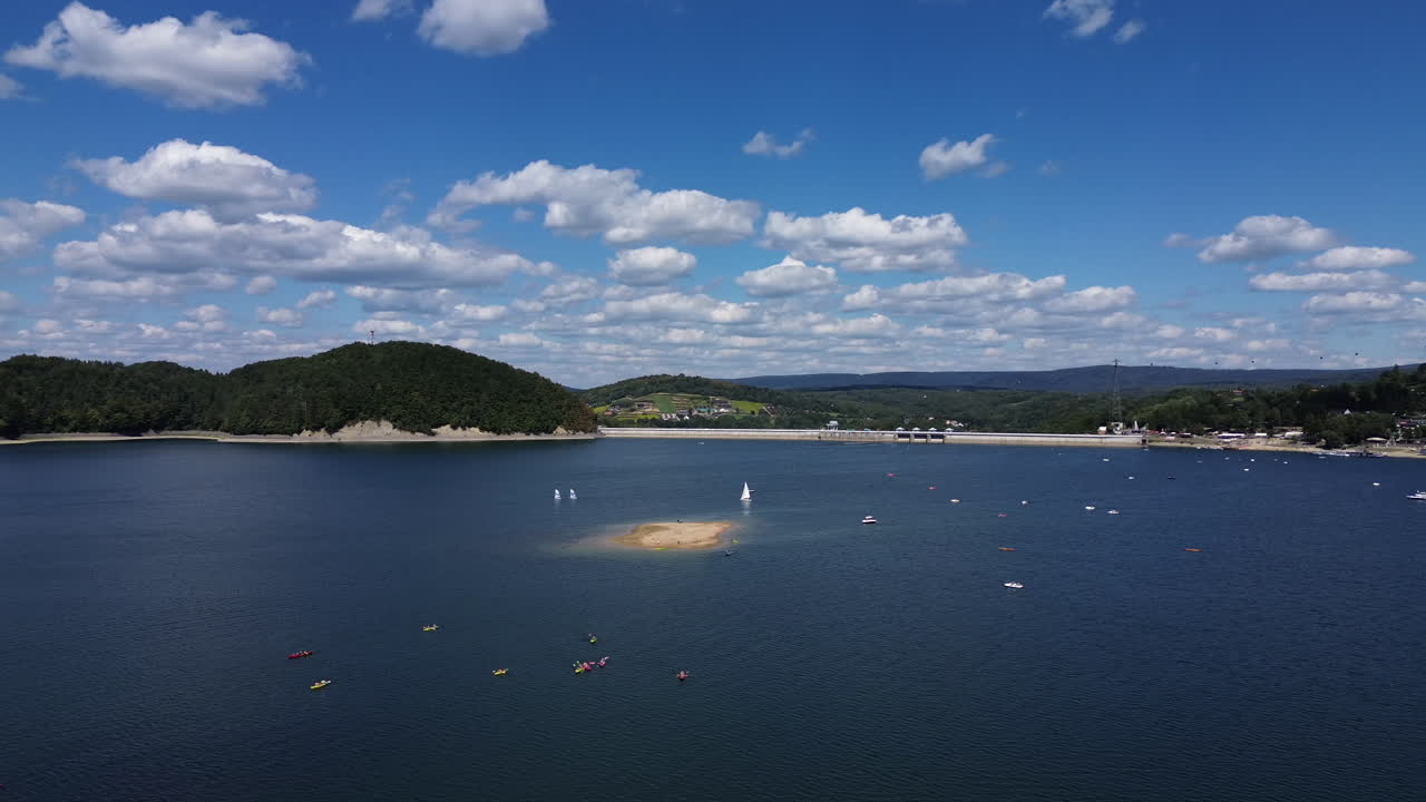 Aerial panning shot of kayaks and sailboats on calm lake with big water dam under a bright blue sky with scattered clouds. Solina, Poland