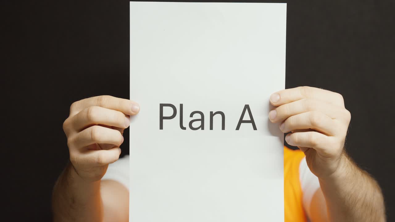 A man in an orange safety vest holds up a paper labeled “Plan A” in front of a black background, representing decision-making and planning at work