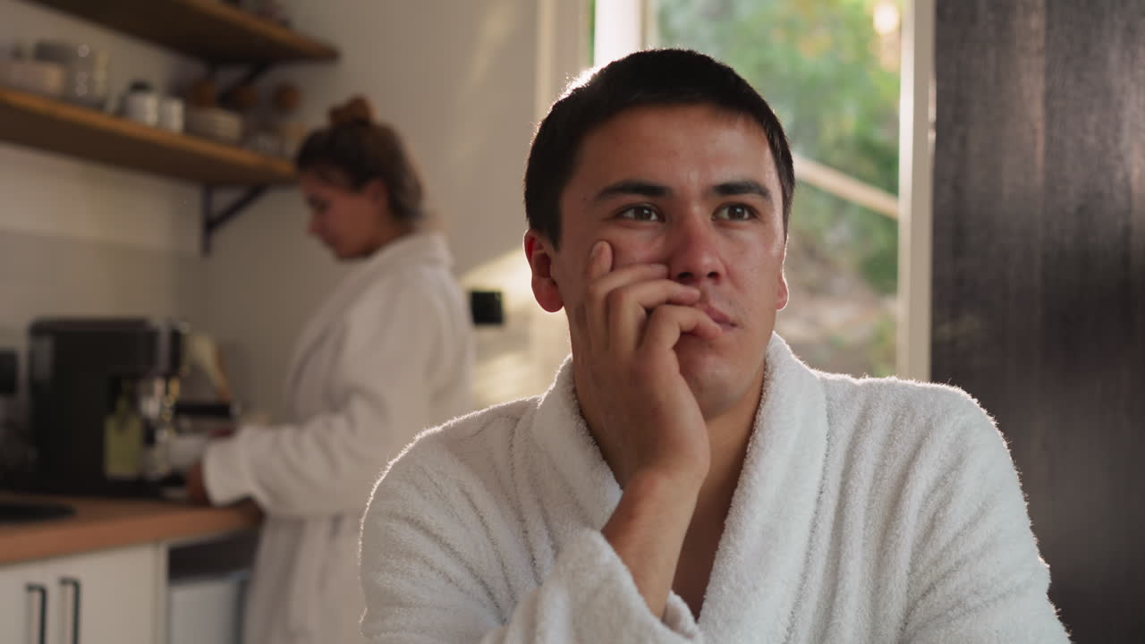 부인이 부에서 커피를 만드는 것을 기다리는 남자: 부드러운 욕조를 입은 사려 깊은 남편은 테이블에 앉아 있고 여자는 아침에 집에서 아침 식사를 요리합니다.