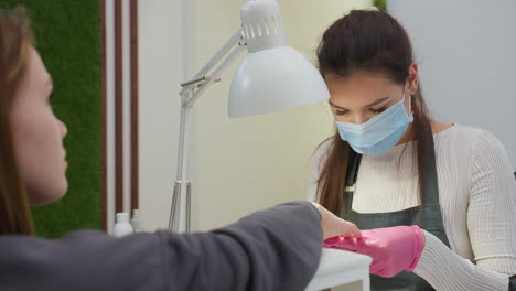 nail technician wearing mask and pink gloves filing fingernails of client sitting across desk near green wall, with client attentively observing process under bright salon lighting during session