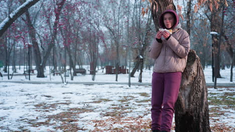 joven con traje de invierno descansando en un árbol comiendo algo en un parque al aire libre durante el invierno, reflejo de luz del fondo, disfrutando de la comida en la naturaleza, momento de relajación en clima frío