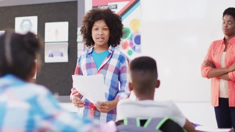 Diverse-female-teacher-and-happy-schoolchildren-at-desks-reciting-in-school-classroom