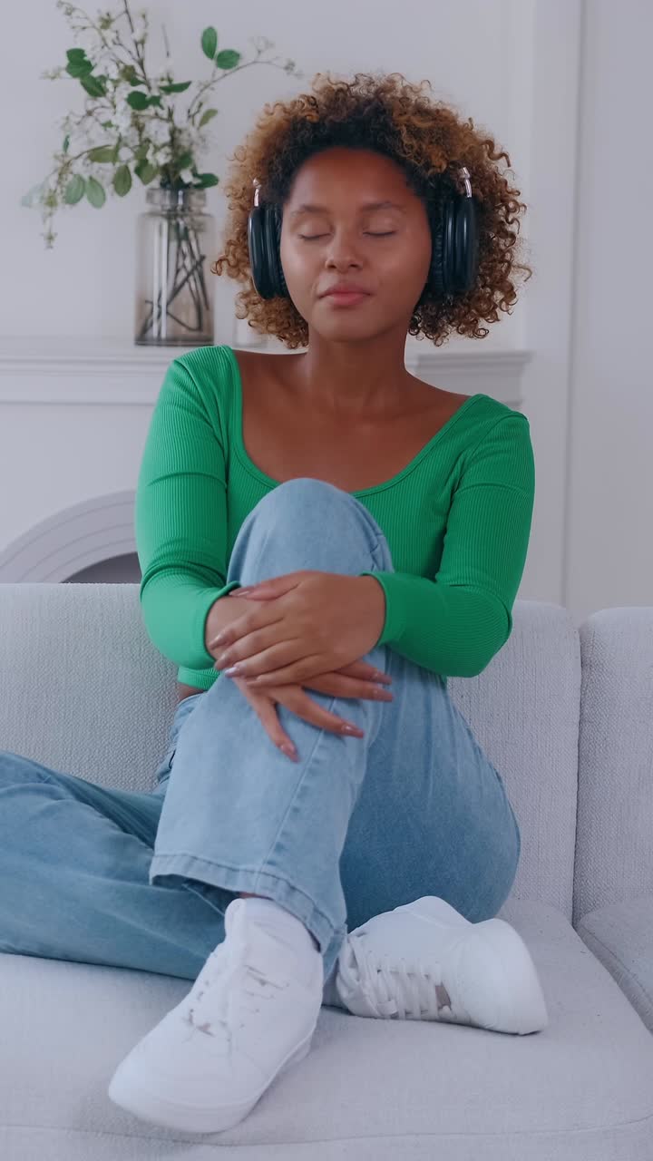Young woman wearing headphones sits on sofa with eyes closed and listening to
