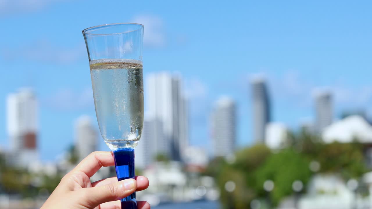 A hand raises a champagne glass filled with sparkling wine against a bright, sunlit city skyline. Shallow depth of field, steady camera, vibrant daylight
