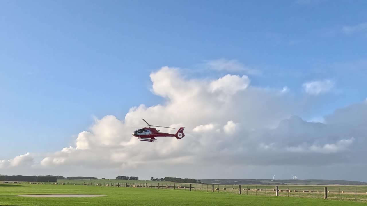 En rød helikopter nærmer sig en grøn mark og hangar for landing under en skyet himmel