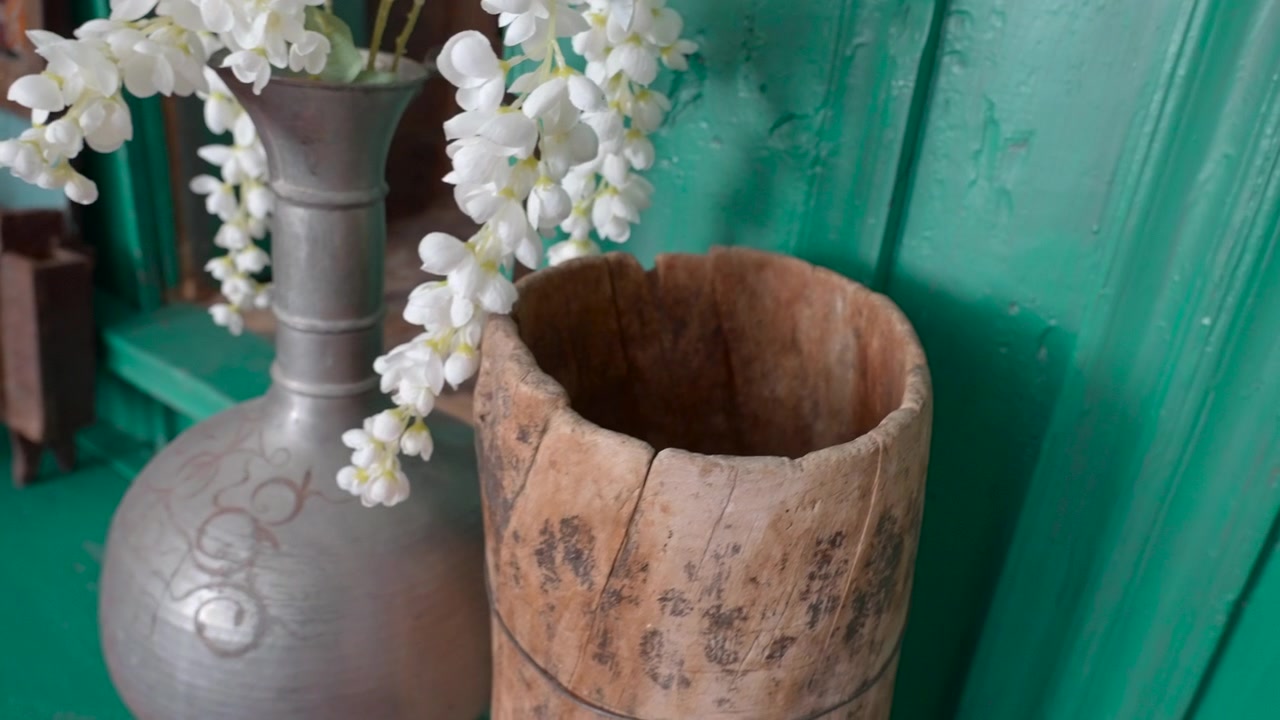 Vista decorativa ravvicinata di fiori artificiali bianchi drappeggiati su un alto vaso in ceramica grigia accanto a un contenitore in legno usurato su una parete dipinta di un vivido color ottanio, con luce soffusa, texture ricca e una calma atmosfera artigianale