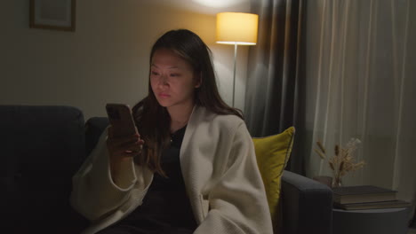 Woman-Spending-Evening-At-Home-Sitting-On-Sofa-With-Mobile-Phone-Scrolling-Through-Internet-Or-Social-Media-1