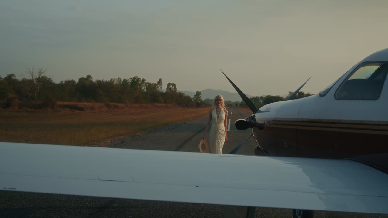 Woman in a stylish dress walking on an airport runway towards a private jet at sunset.