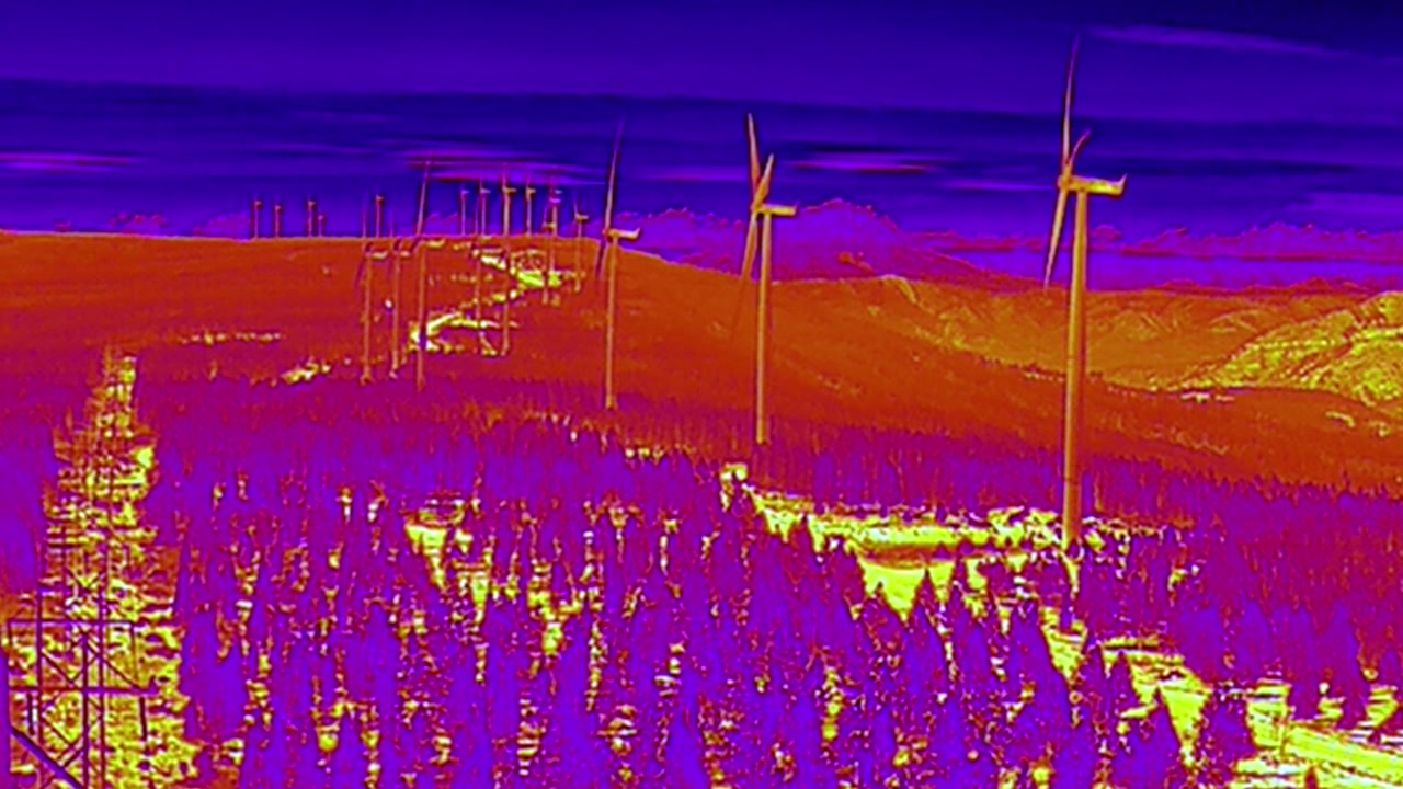 Termisk bildebehandling avslører vindturbiner på en skogkledd åsrygg i skumringen. Ideell for energidokumentarer eller miljøstudier