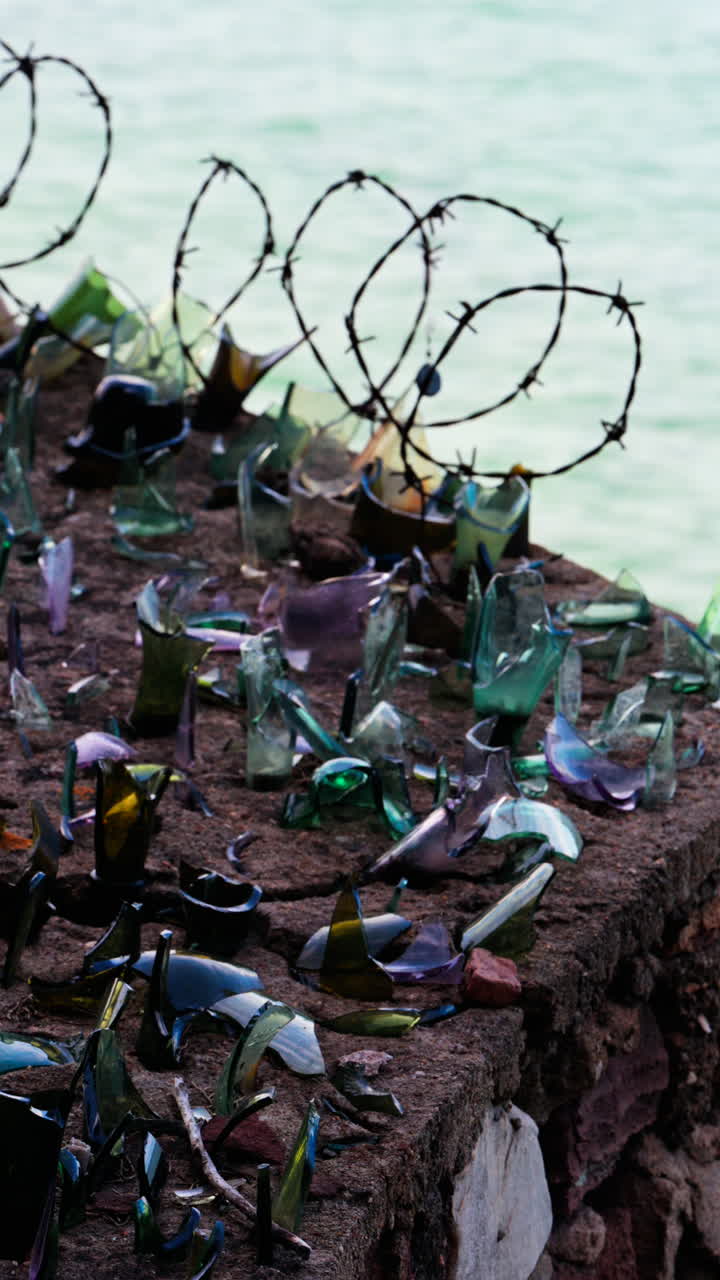 Close up of multiple broken glass shards and barbed wire loops along the edge of a brick wall with a blurred view of the sea. Vertical