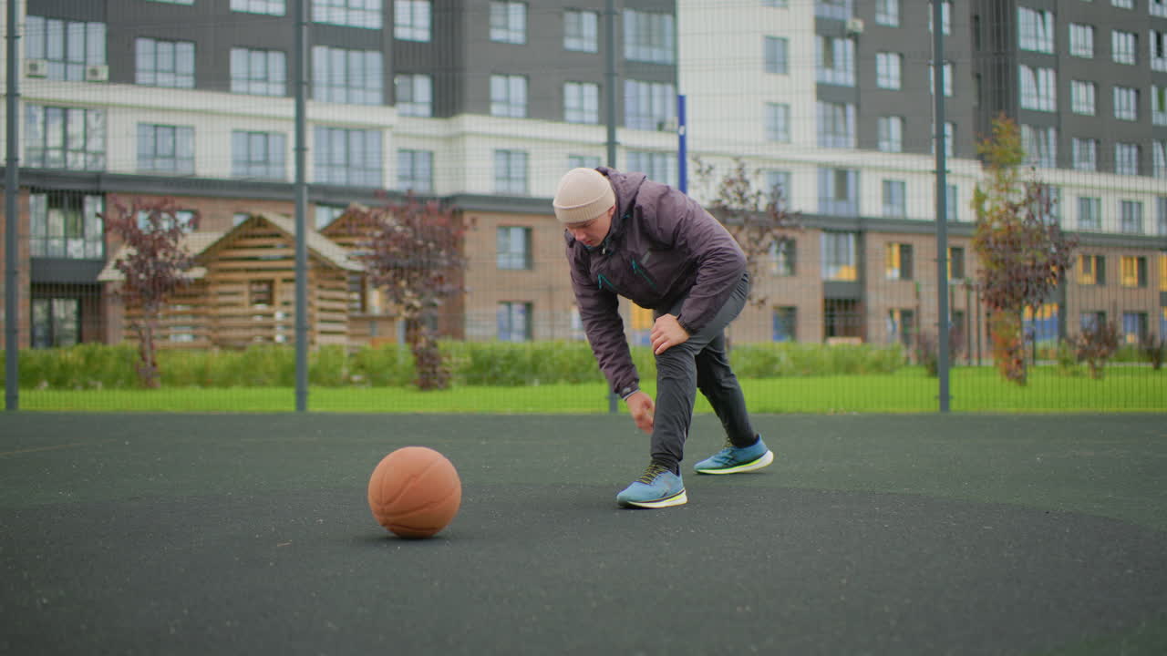 Senior caucasian man tying shoes beside ball, kneeling to adjust laces on outdoor court, closeup on steady hands and sneakers, apartment buildings and fence in background, cool cloudy morning, pregame