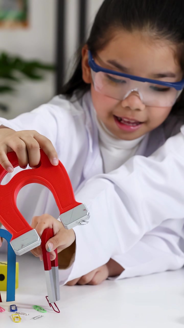 Siswa muda mengenakan jas lab menggunakan magnet tapal kuda untuk mengambil klip kertas selama pelajaran sains.