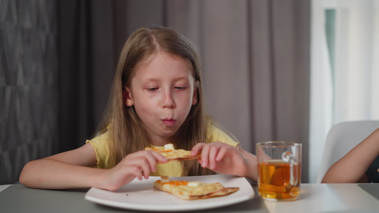 little girl wearing yellow shirt enjoying slice of pizza at dining table with glass of berry juice beside her while another person is partially visible
