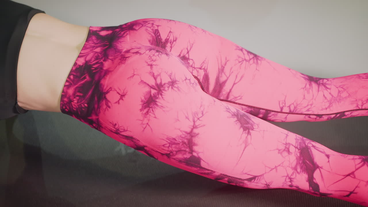 donna in leggings rosa che fa plank su un tappetino, primo piano di torso e gambe che mostrano l'attivazione del core, supporto sugli avambracci, respiro regolare, allineamento dell'anca controllato, illuminazione da studio, allenamento casalingo casual, fitness