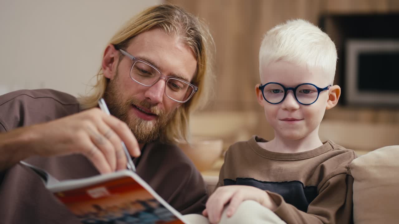 primer plano: un hombre rubio con barba y gafas ayuda a su pequeño hijo albino de cabello blanco con gafas redondas azules a hacer su tarea y le lee lo que está escrito en un cuaderno. sentado en el sofá de un apartamento moderno por la noche