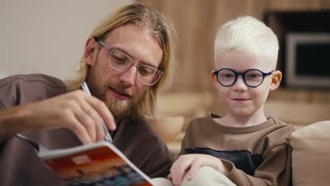 Close-up:-a-blond-man-with-a-beard-and-glasses-helps-his-little-albino-son-with-white-hair-in-blue-round-glasses-to-do-his-homework-and-reads-for-him-what-is-written-in-a-notebook.-Sitting-on-the-sofa-in-a-modern-apartment-in-the-evening