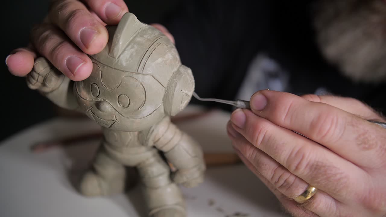 A close-up of hands sculpting a small cartoonish clay figure with a helmet and big eyes. The artist uses a carving tool to add fine details, with the figure standing on a circular base.