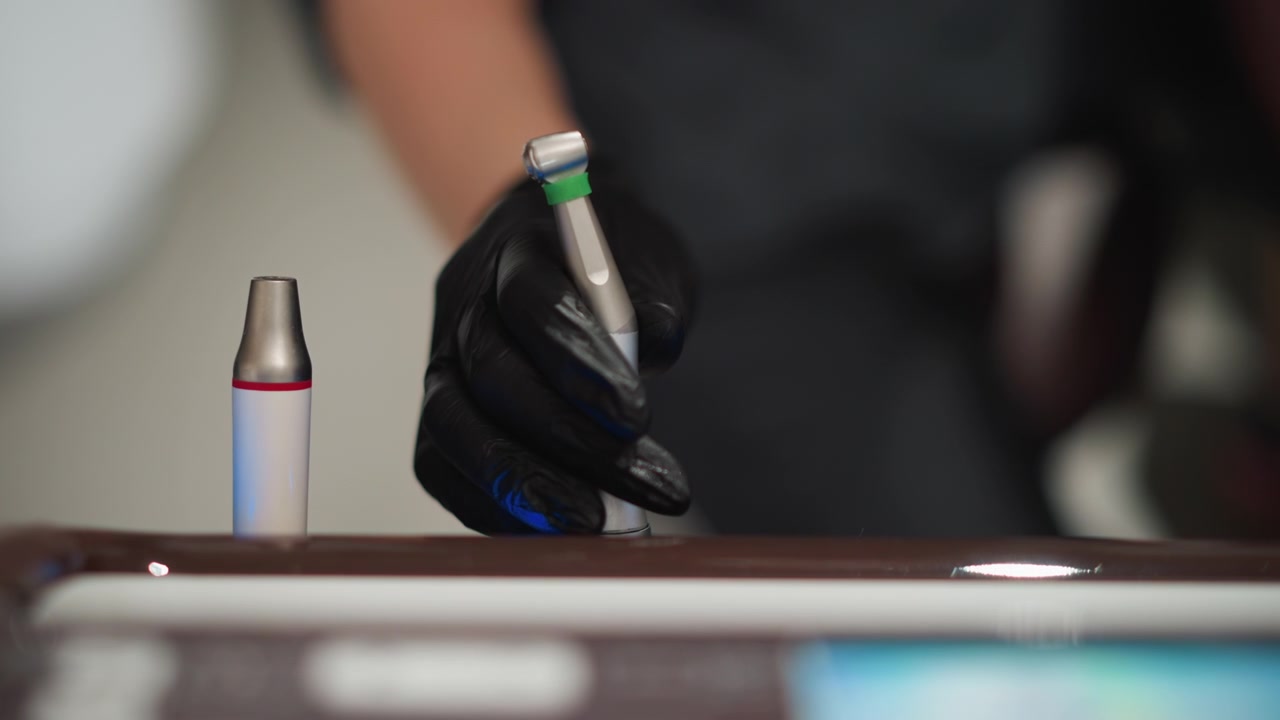 Primo piano di mano guantata che afferra un manipolo dentale sul bancone. Strumenti dentali, inclusi manipoli e siringa, visibili con illuminazione soffusa che evidenzia le texture metalliche. Sfondo