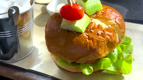 a close-up of a gourmet burger topped with avocado and cherry tomato, served with fries in a metal bucket.