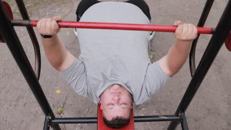 a fat man in a gray tshirt lifts weights while lying on an outdoor exercise machine a fat man in a gray tshirt lifts weights while lying on an outdoor exercise machine struggle with excess weight lifting the bar
