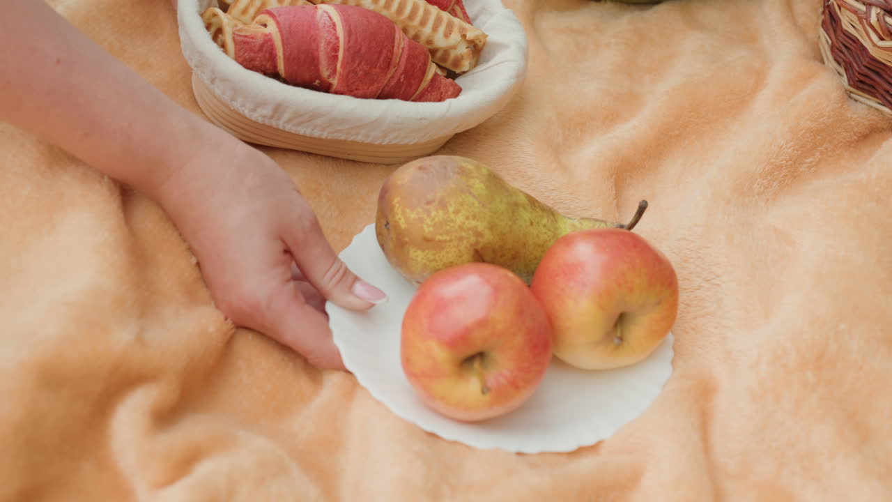 Mano che sistema delle mele su una coperta, piatto bianco con pera, cesto di vimini con dolcetti, primo piano di una mano femminile che prepara un accogliente picnic autunnale su un morbido tessuto beige, calda luce solare e atmosfera rilassata.
