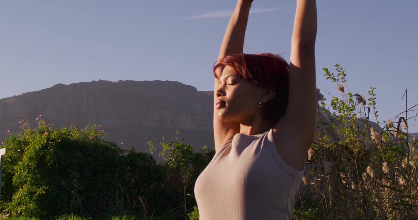 Donna afroamericana con il sole che le riscalda il viso, che si allunga all'aperto alzando le braccia, in canottiera, con spazio per il testo.