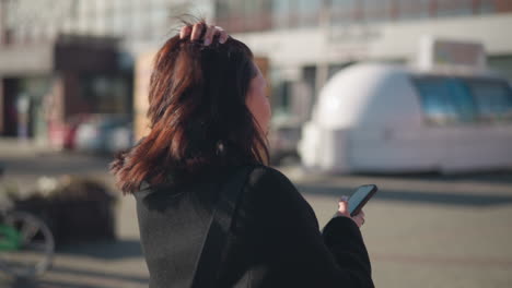 woman walking outdoors adjusting her hair while holding a phone, briefly looking around before focusing back on her device, background features modern urban architecture and green bicycle