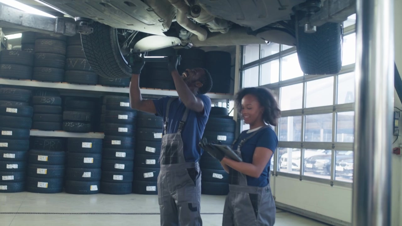 Diverse pair of smiling mechanics have fun while working together inspecting the underside of a