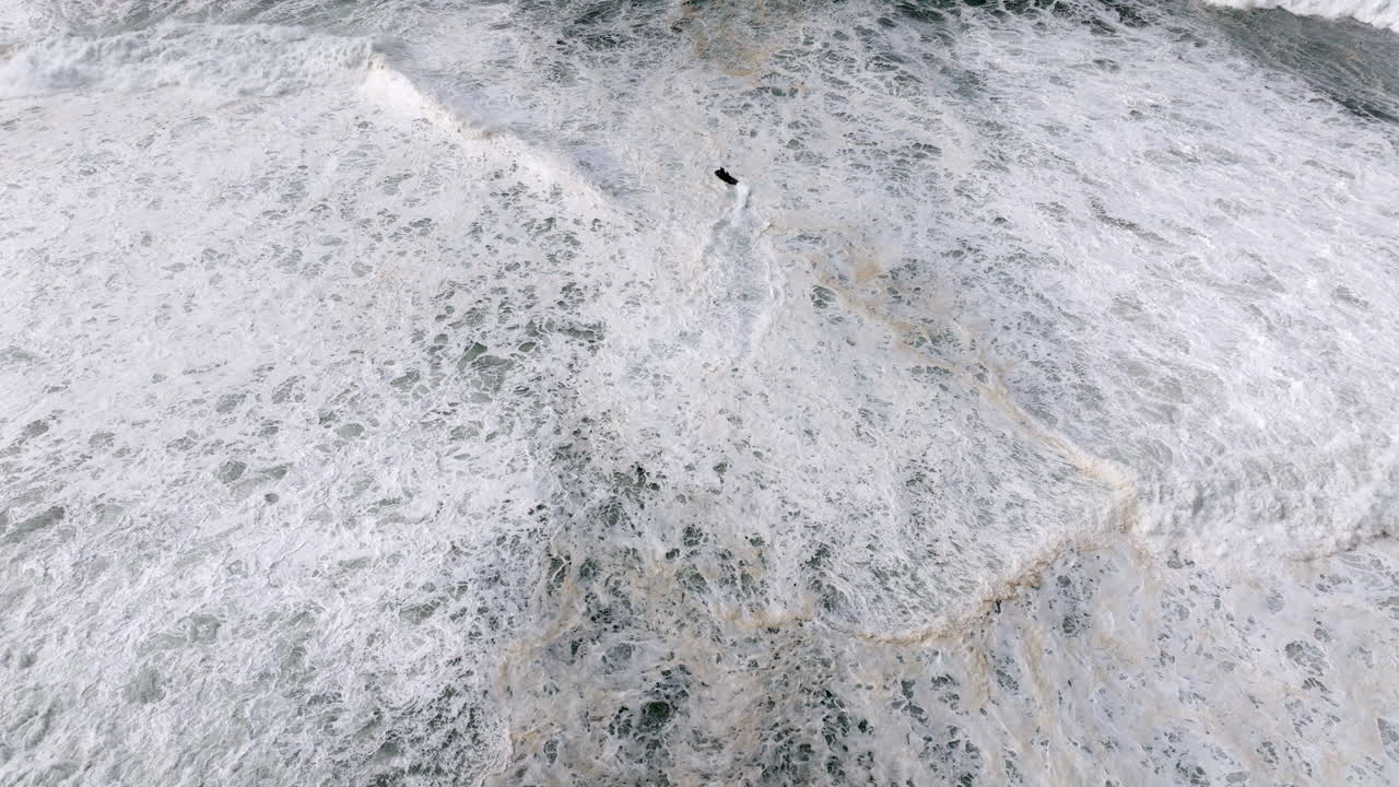 Jet ski team rider navigating giant storm waves at Nazare, Portugal. Professional big wave tow-in surfing location. Top-down aerial drone establishing shot, with white water and big waves breaking