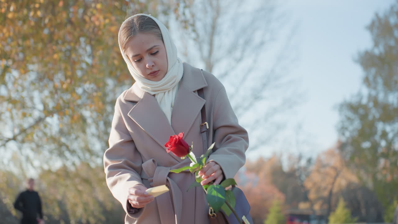 Donna bianca che sistema una rosa rossa vicino a un lampione in un parco autunnale, con l'atteggiamento pensieroso di una studentessa, che taglia lo stelo e ispeziona i petali; cappotto leggero e sciarpa, sfondo di un memoriale urbano, luce solare soffusa e alberi spogli.