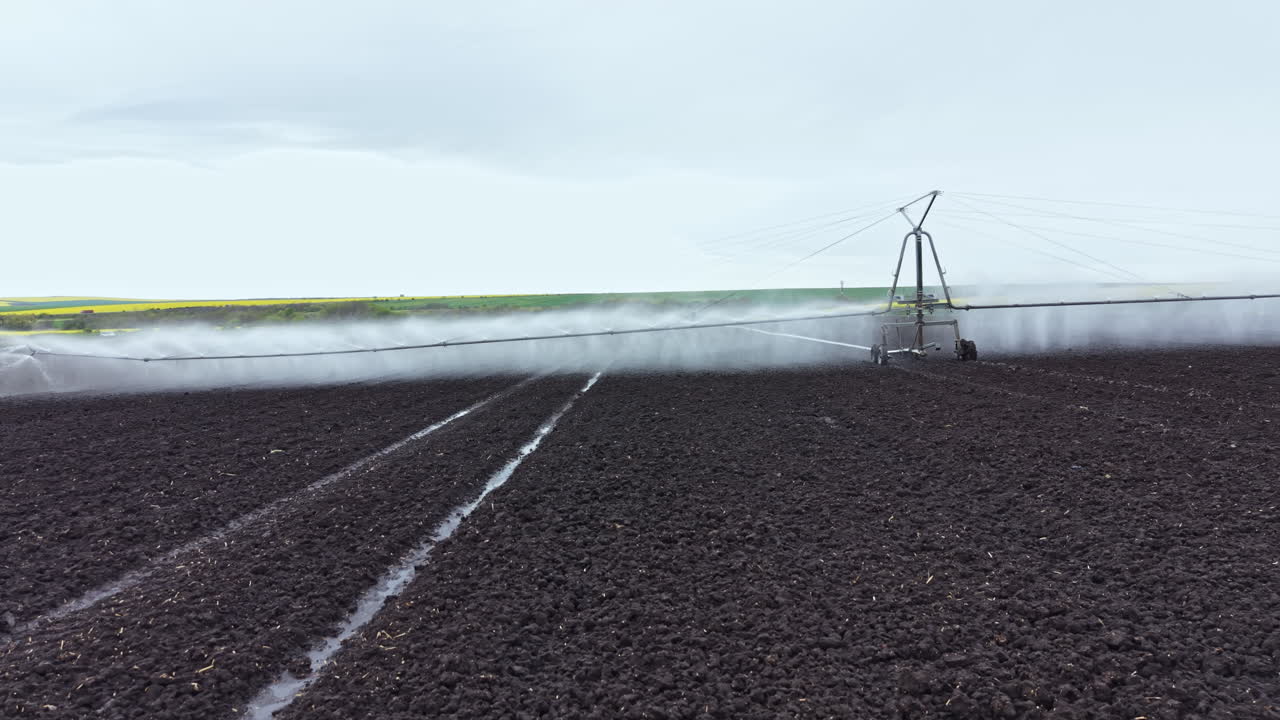 Campo agricolo con impianto di irrigazione che spruzza acqua su terreno arido sotto un cielo nuvoloso nelle prime ore del mattino in un paesaggio rurale.