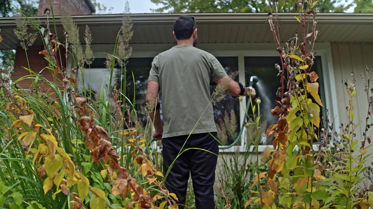 Man washes a large house window with a garden hose and brush. He stands in a garden filled with plants in full fall colors