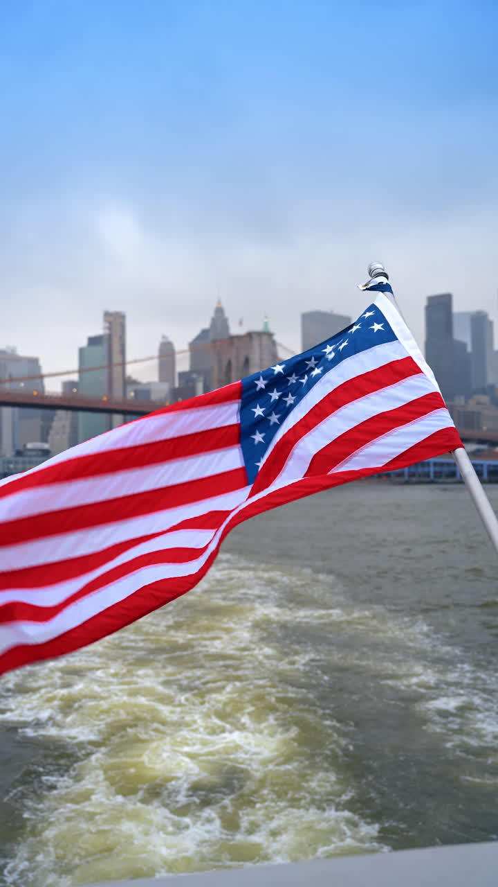 Bandiera americana che sventola su un'imbarcazione con lo sfondo del Ponte di Brooklyn. Bandiera degli Stati Uniti fissata a un'imbarcazione che naviga sull'East River, con lo skyline di New York City e il ponte alle spalle.