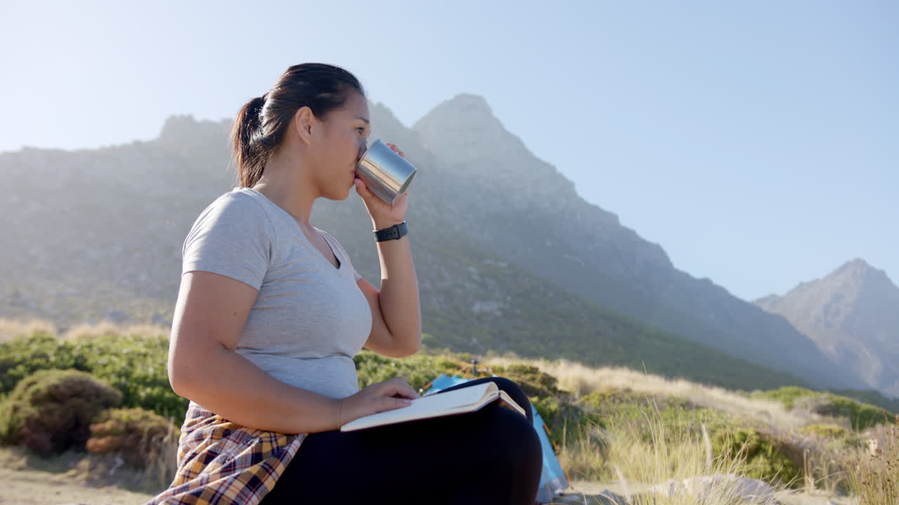 Donna che beve da una tazza, seduta su un sentiero di montagna con un quaderno.