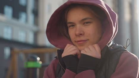 portrait of cheerful outdoor trainer covering face with maroon hoodie and black vest, bright sun glare, playful pose, confident gaze shining through hood during park workout session